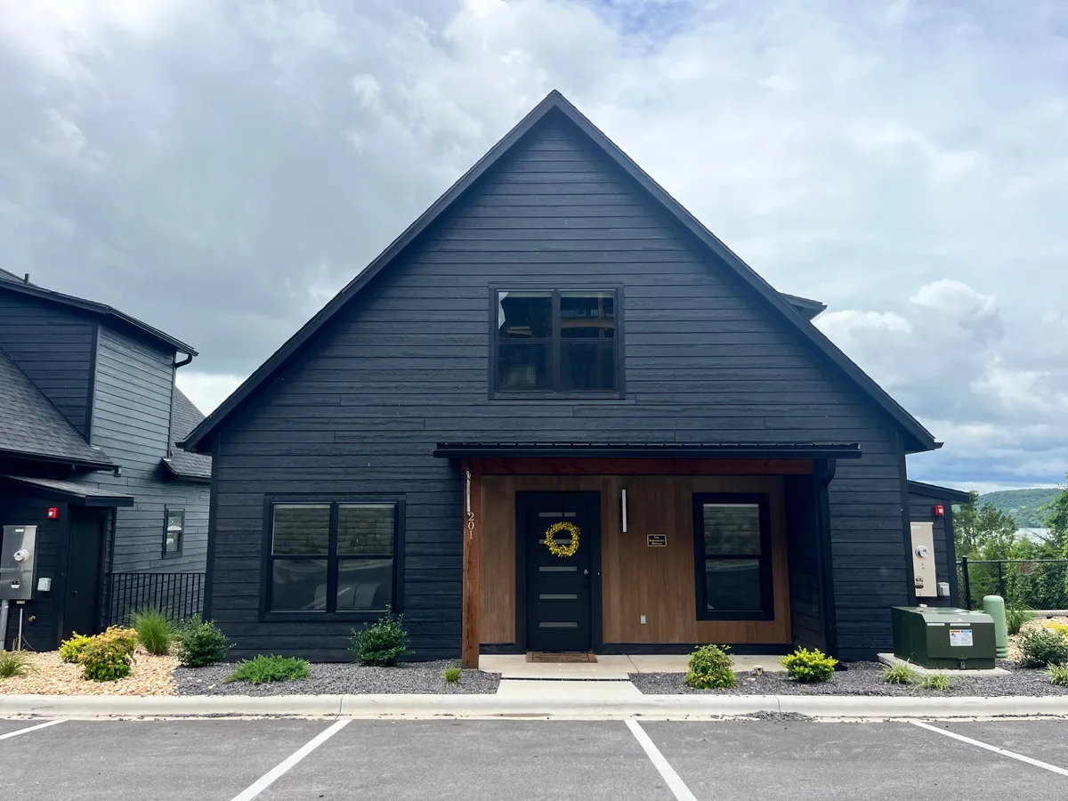The exterior of a contemporary home with a dark wooden facade, a cheerful yellow wreath on the front door, large windows, and a beautifully landscaped garden. The spacious porch area invites outdoor relaxation and hints at ample room for pets to explore and enjoy the fresh air alongside their owners.