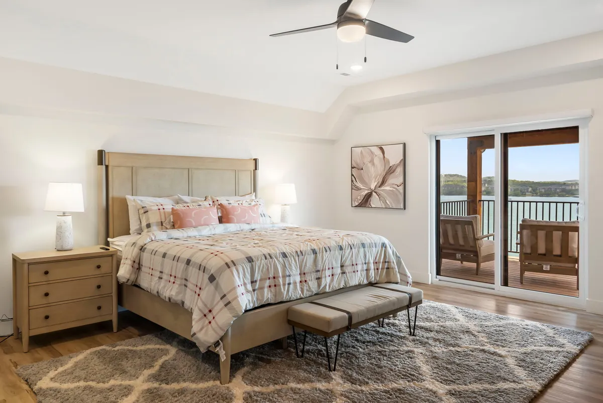 A serene bedroom featuring a comfortable bed with soft bedding, decorative artwork above the headboard, a nightstand with a lamp, and sliding glass doors revealing a scenic view. Light fills the room, creating an inviting atmosphere for rest and relaxation.