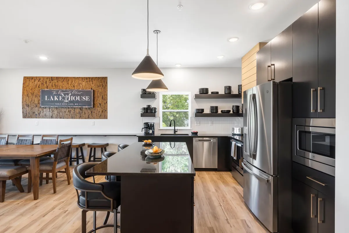 A bright and welcoming kitchen and dining space with a prominent kitchen island, dining table, 'Lake House' sign, and black cabinetry. Large windows bathe the area in natural light and provide glimpses of the lakeside, echoing the narrative of lakeside living and shared culinary experiences.