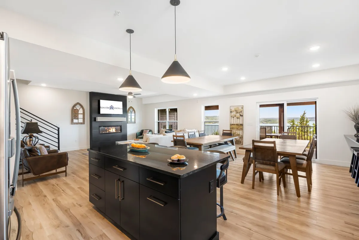 This image captures a spacious, sun-filled open-concept interior that seamlessly integrates the kitchen, dining, and living areas. The kitchen island is central, offering generous counter space and modern appliances, while the dining area and living room extend towards expansive windows showcasing a beautiful lake view. Warm, inviting tones and comfortable furnishings set the atmosphere for gatherings and shared meals.
