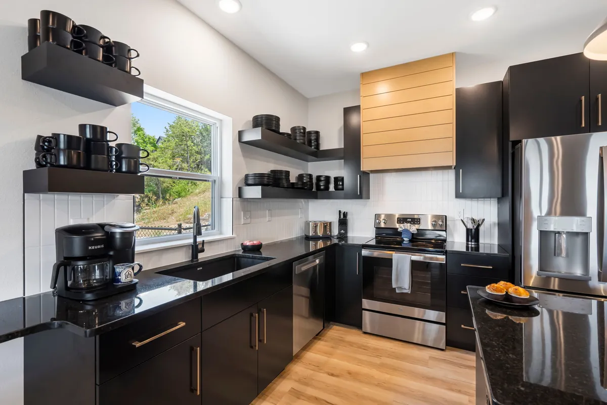 A spacious, contemporary kitchen with sleek black cabinetry, shiny stainless steel appliances, and open shelving. A large window brings in natural light, highlighting the pristine and luxurious setting.