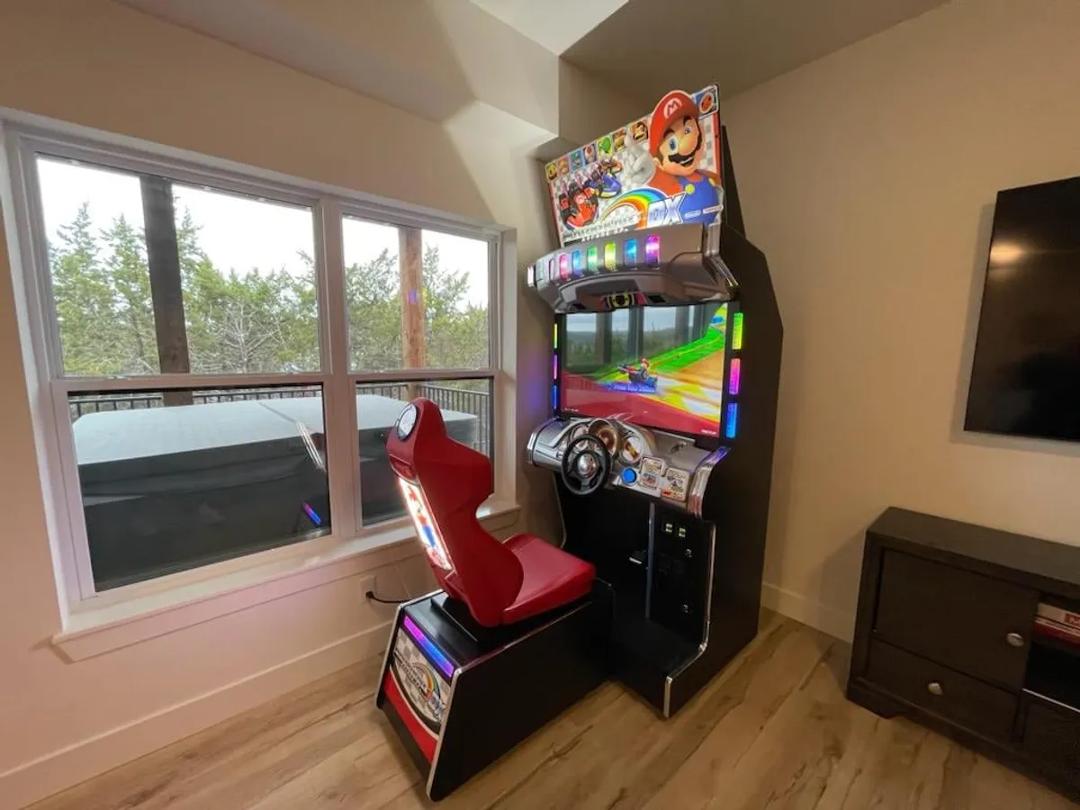 A bright and energetic game room anchored by an arcade racing machine, large windows letting in natural light, and wooden flooring. The setup invites friendly competition and nostalgic fun for families and adults alike.