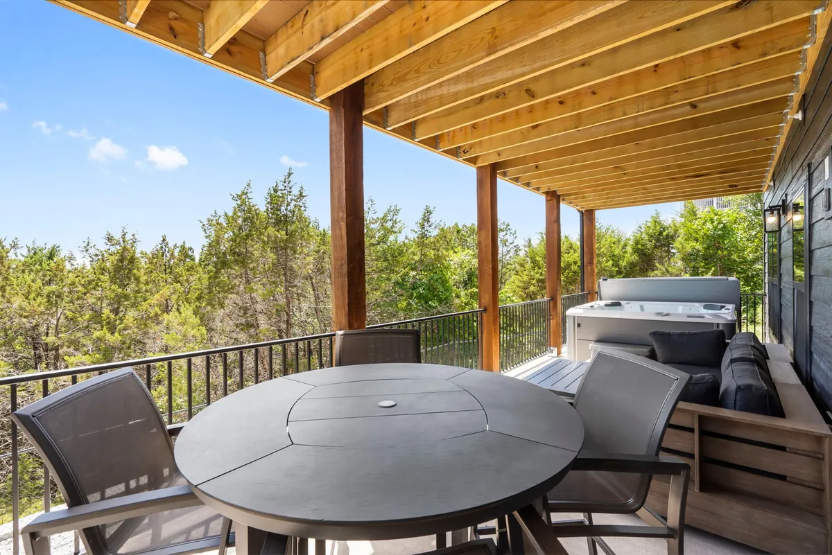 A spacious outdoor deck featuring a dining table, comfortable seating area, a bubbling hot tub, and wooden beams, all surrounded by lush greenery. The deck is set up for both relaxation and social gatherings, with plenty of space for lounging or enjoying meals outdoors.