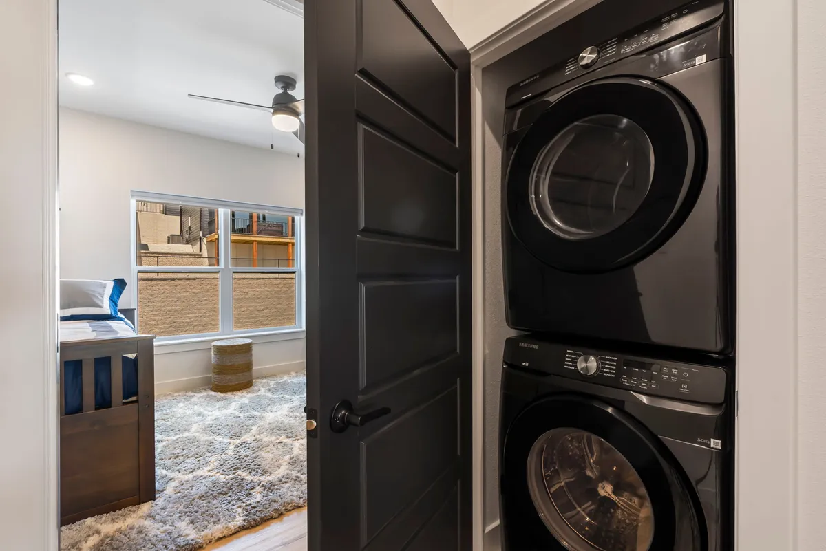 A convenient in-home laundry area with a stacked washer and dryer, visible bedroom windows, and a textured area rug, blending functionality with comfort and natural light.