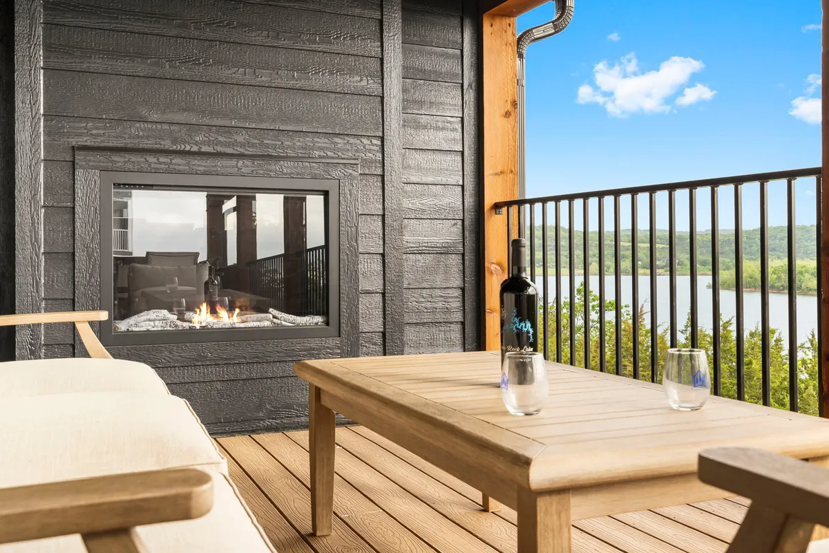 An inviting outdoor patio with a cozy fireplace, a coffee table set with wine and glasses, and a sweeping lake view beyond. The scene is set for relaxing evenings, group gatherings, and enjoying sunsets or stargazing from the comfort of the deck.