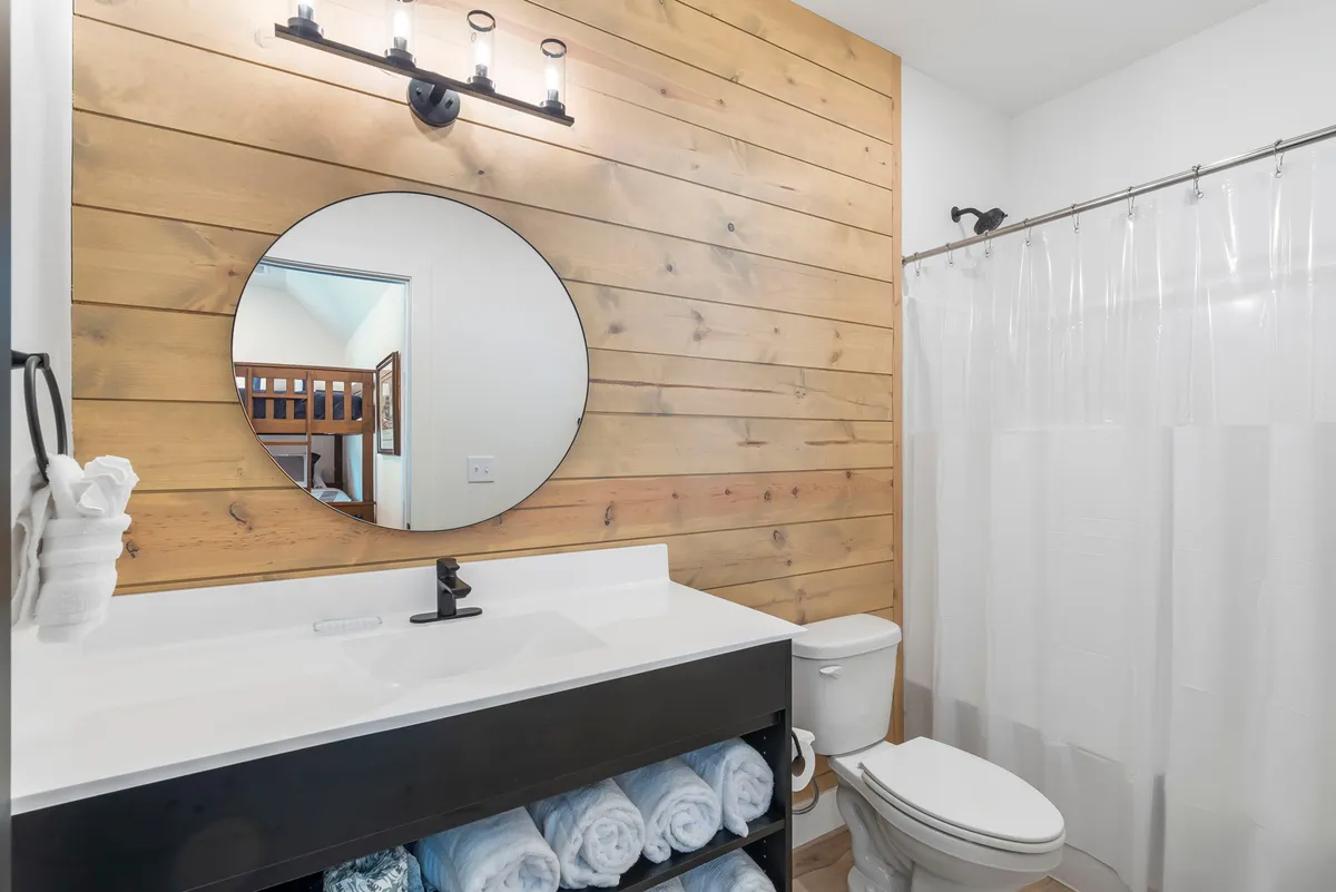 A modern bathroom scene with a circular mirror, black vanity, wooden accent wall, soft lighting, and neatly arranged towels. The clean and cozy setting is perfect for refreshing after a long day, reflecting the thoughtfully designed ensuite and spacious bathrooms.
