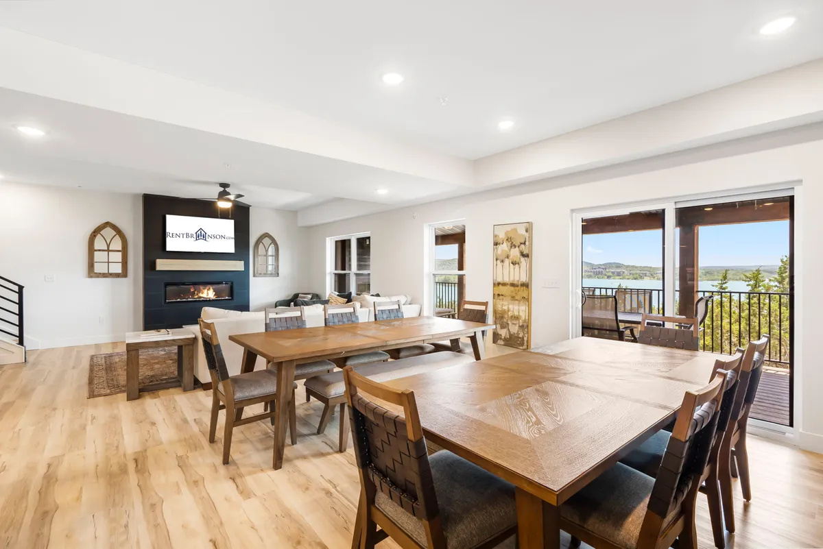 A welcoming interior scene featuring a large wooden dining table with ample seating for groups, a cozy stone fireplace, and sliding glass doors that open to scenic outdoor views. The textured wood accents and comfortable seating create a sense of warmth and togetherness, ideal for family meals or celebratory dinners.