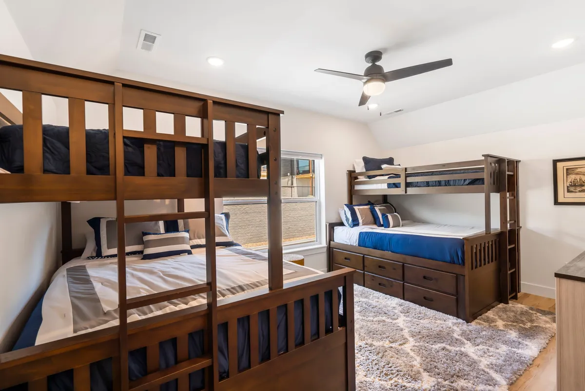 A playful and spacious bunk room filled with natural light. Two sets of bunk beds, a full-size bed with drawers, a colorful decorative rug, and windows create a cheerful and practical space for children or groups. The atmosphere invites play, sleepovers, and family bonding.