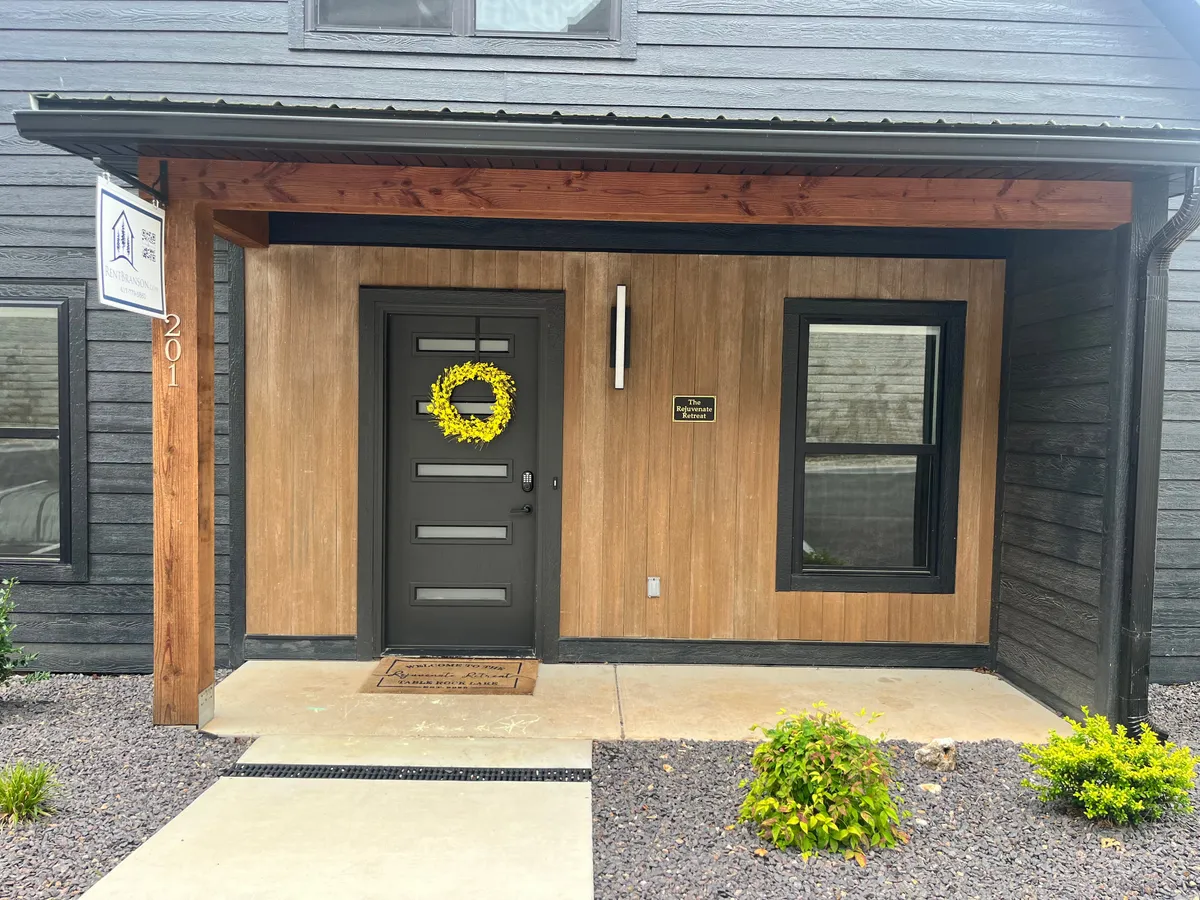 This image presents a charming front porch with a yellow wreath on the door, flanked by landscaped plants and a prominent house number sign. The wooden porch exudes a sense of welcome and care.