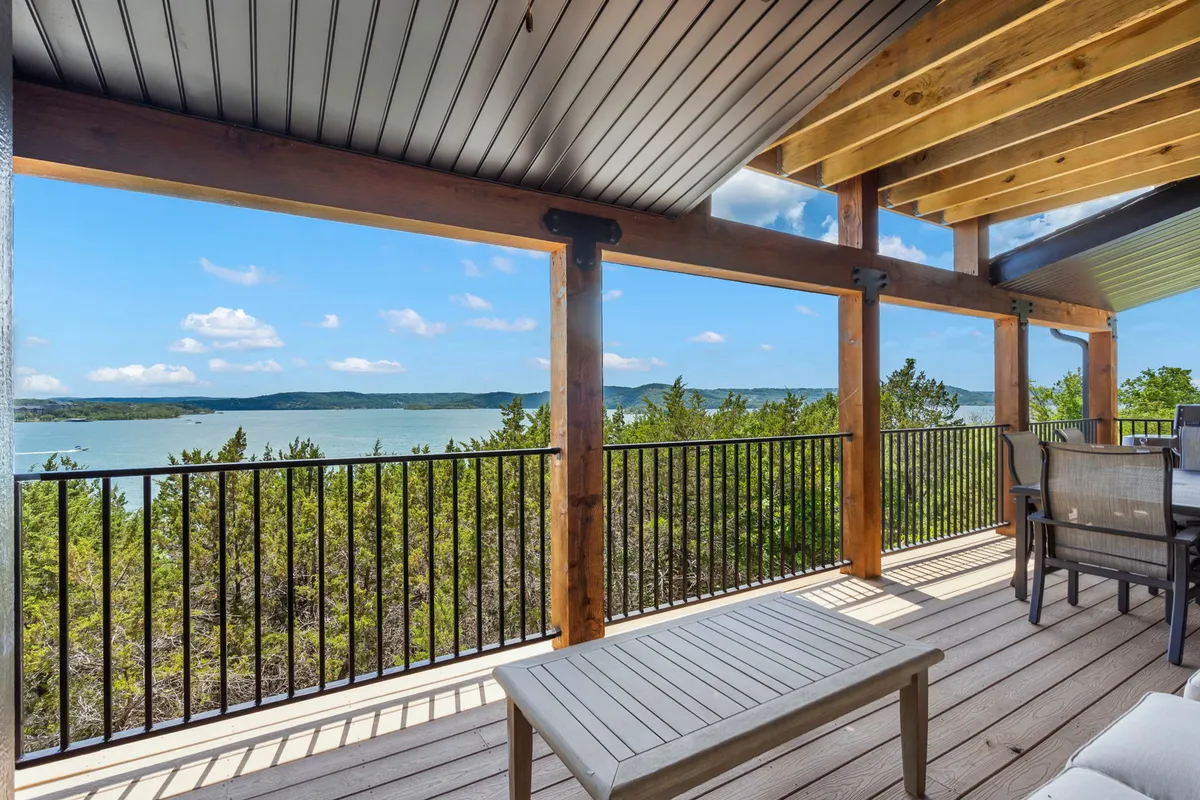 A wooden deck with comfortable outdoor furniture overlooks calm lake waters and a glowing sunset sky, framed by a rustic railing. The scene invites guests to enjoy a morning coffee with a view or unwind at the end of the day.