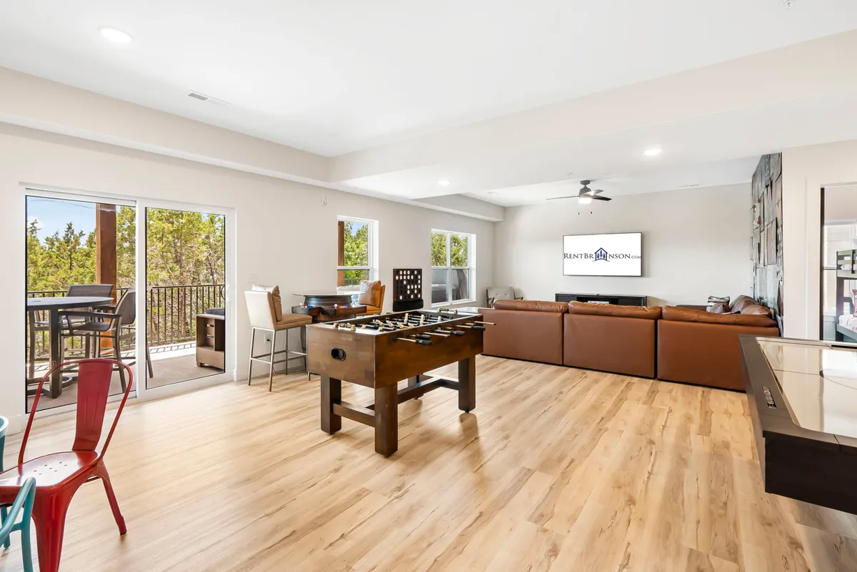 A spacious lower-level living area featuring a foosball table, a large sectional sofa, a TV, and windows with an outdoor view. The setting is relaxed and social, ideal for both competitive games and unwinding after play.